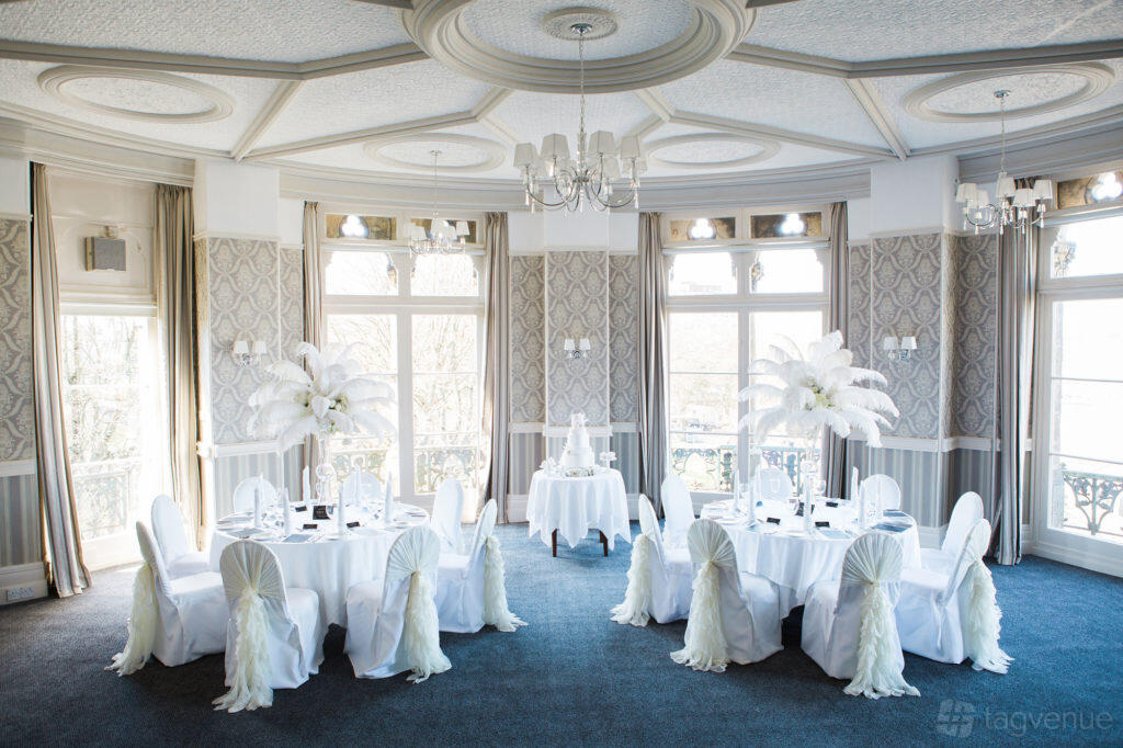 An event space with round tables draped in white linens, feather centerpieces, and large bay windows at The Duke of Cornwall Hotel.