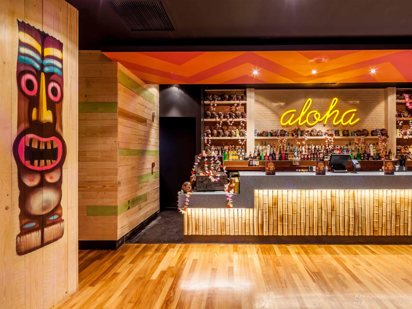 A nightclub bar with bamboo paneling, neon aloha sign, tiki mugs, and a painted mask mural at PRYZM Brighton.
