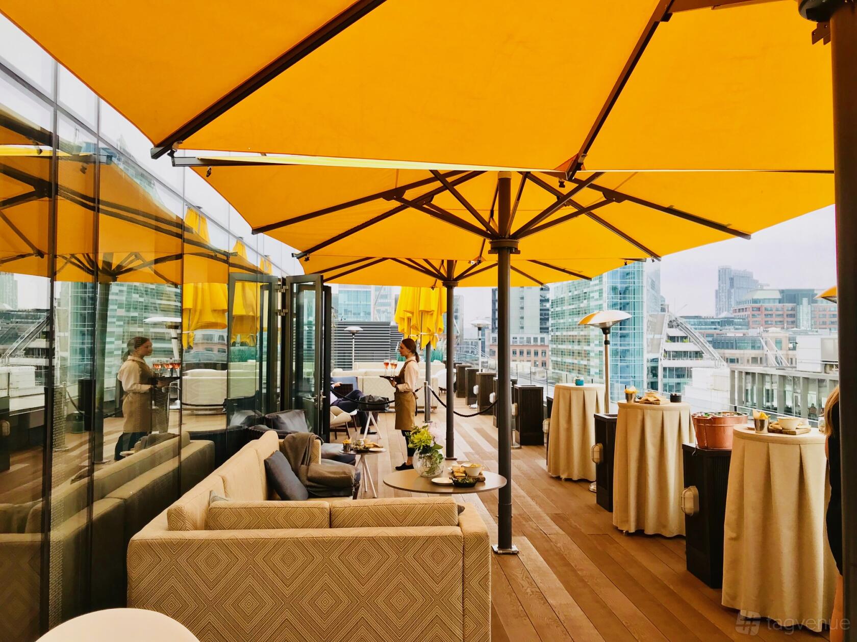 A terrace bar with yellow umbrellas, cushioned seating, cocktail tables, and city views at Aviary Rooftop Bar & Restaurant.