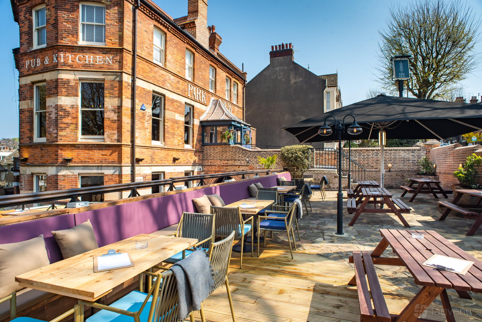A pub terrace with wooden tables, cushioned bench seating, and large umbrellas at The Park View.
