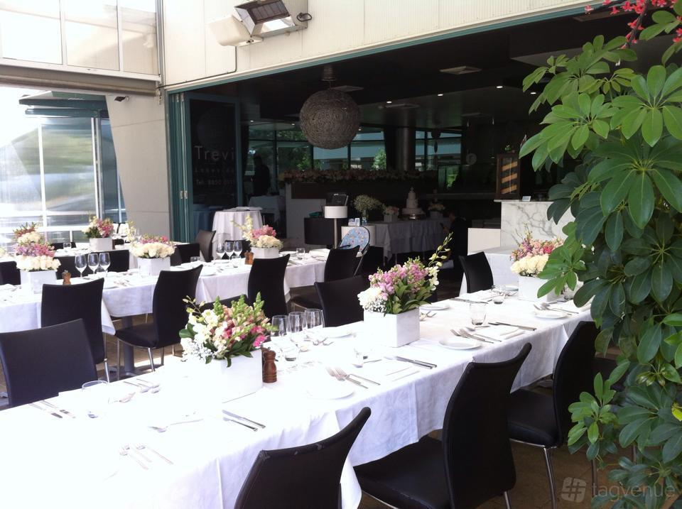 A restaurant with long tables draped in white linens, black chairs, and floral centerpieces at Trevi Lakeside.