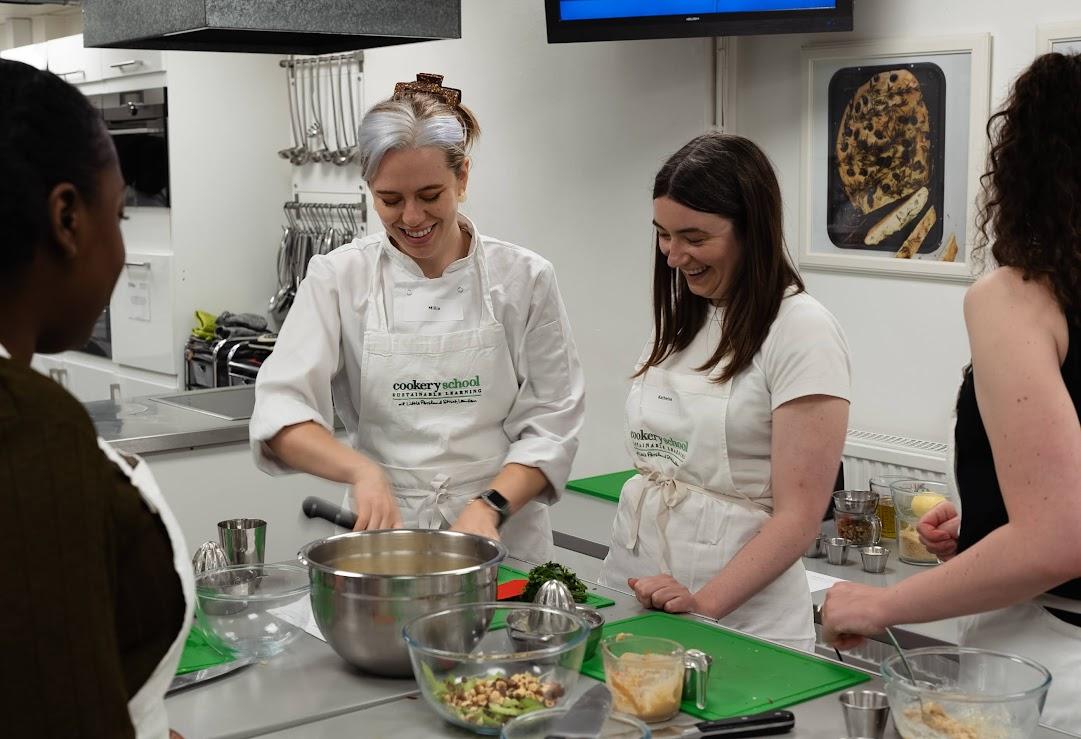 A school kitchen with stainless steel countertops, mixing bowls, and people preparing food at Cookery School at Little Portland St