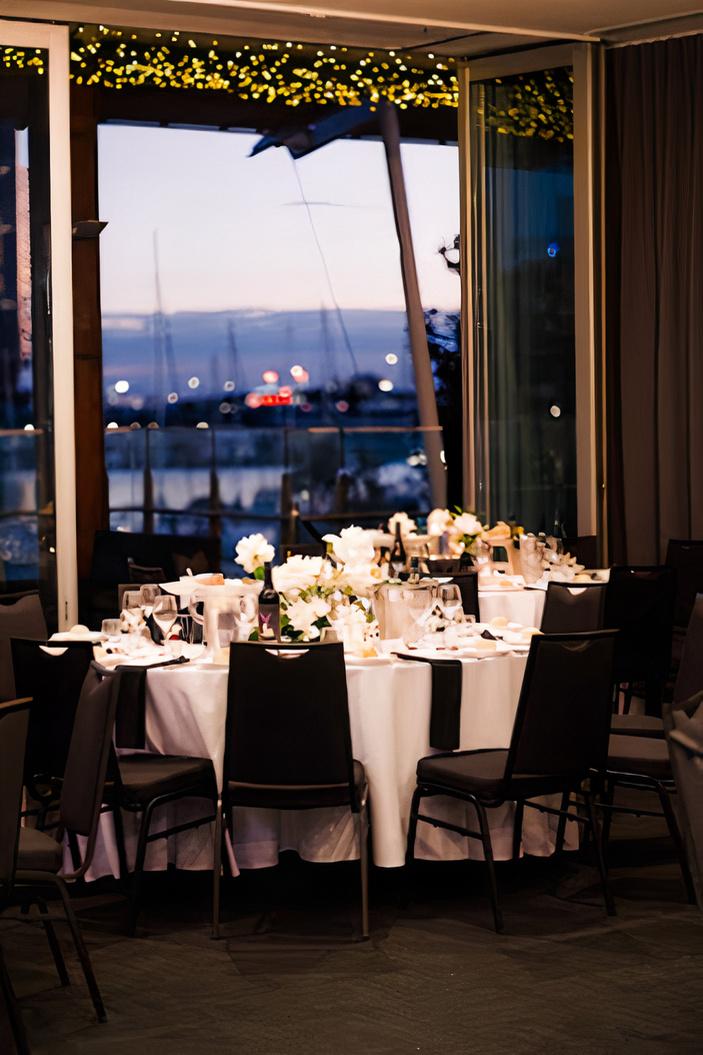 A banquet hall with round tables set with white linens, floral centerpieces, and harbor views at The Promenade Docklands.