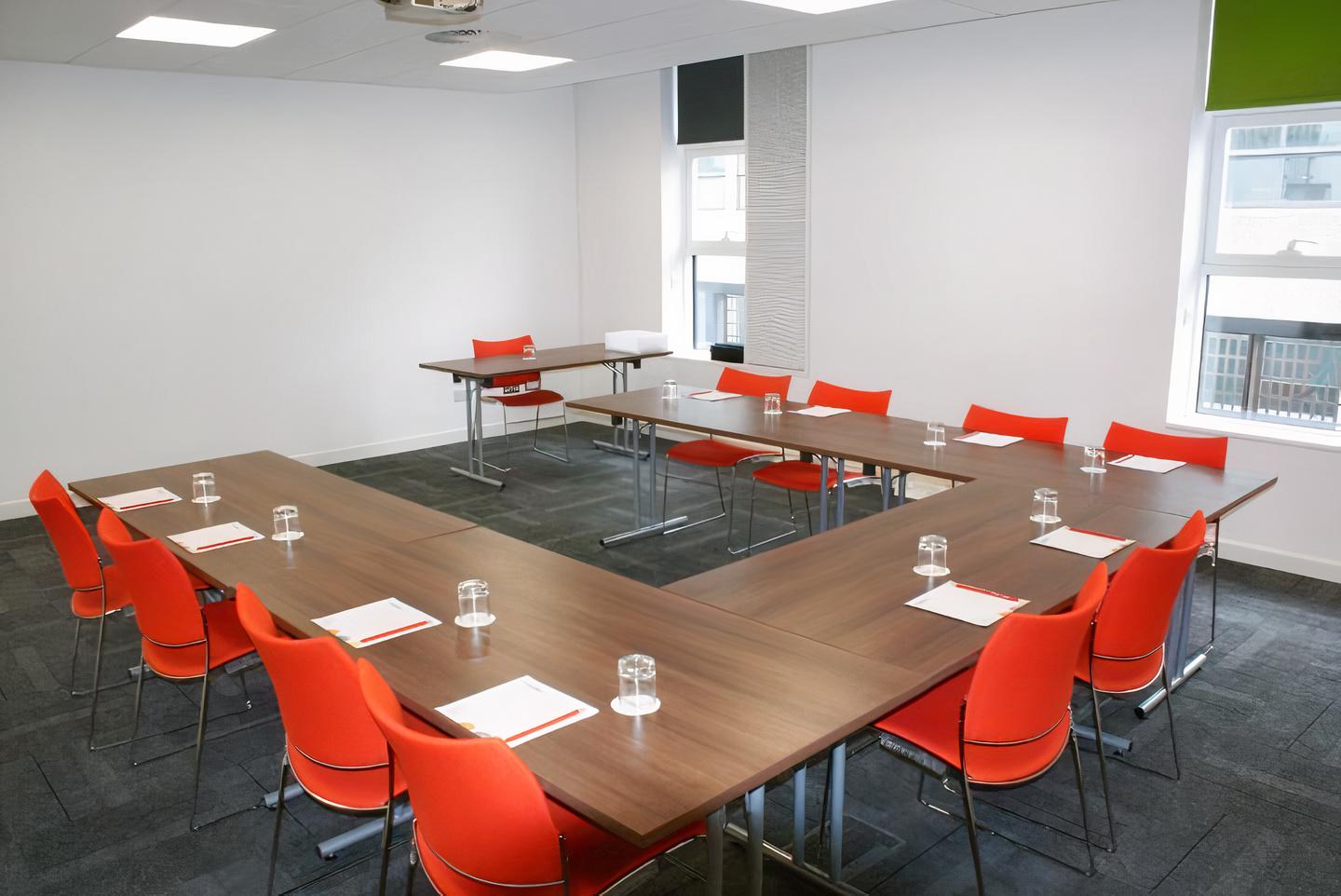 A conference centre meeting room with U-shaped tables, red chairs, and large windows at The Studio Manchester.