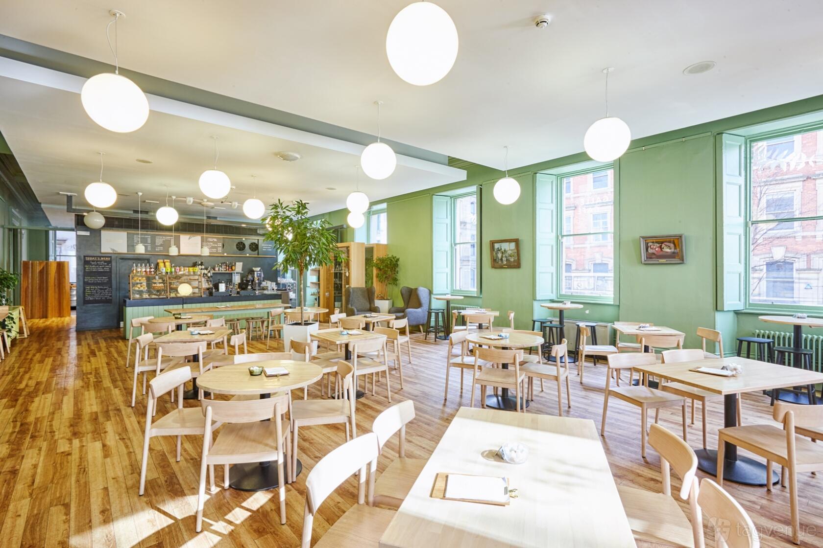 A gallery café with round wooden tables, pale chairs, globe pendant lights, and tall windows at Manchester Art Gallery.