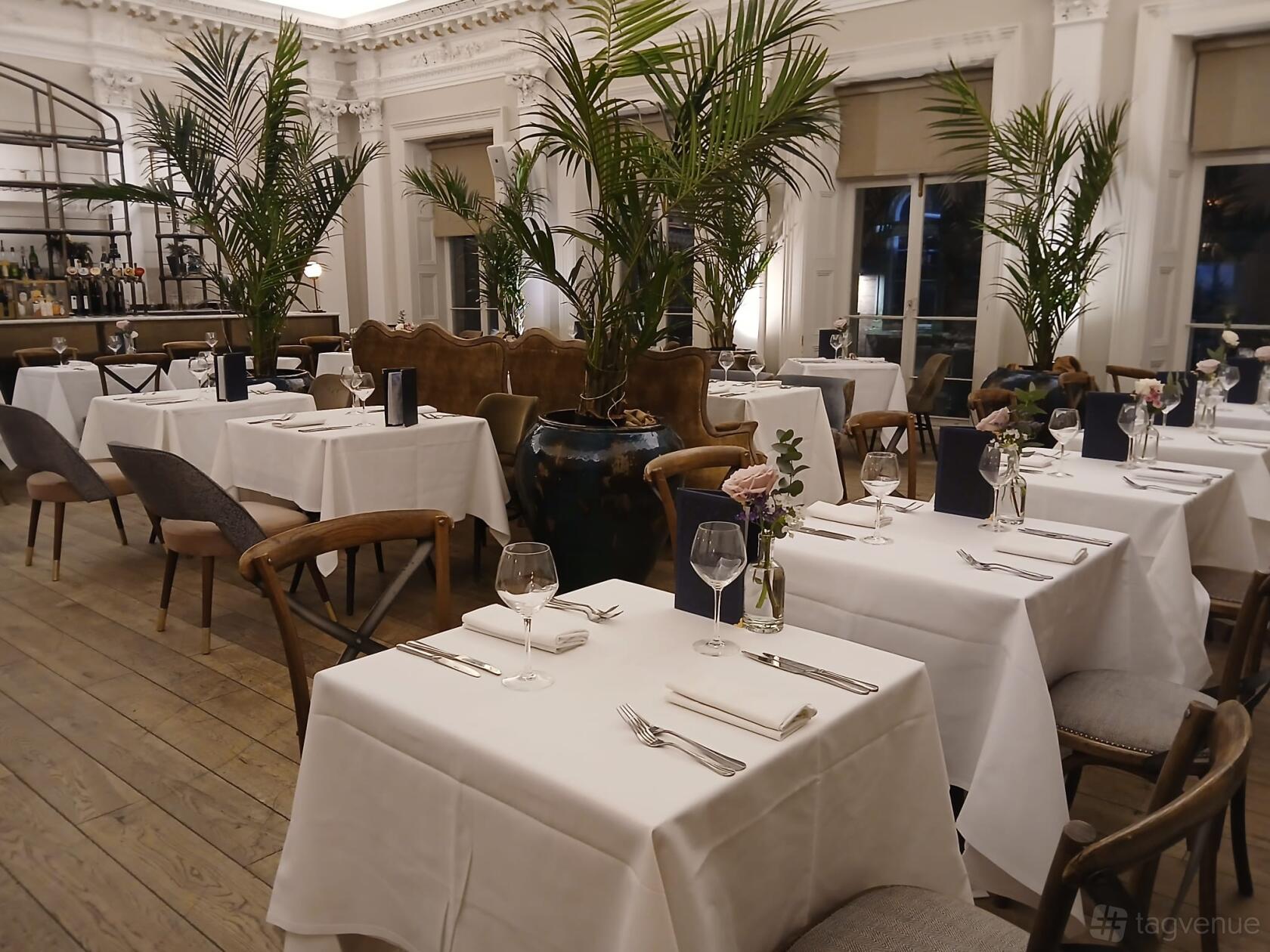 An event space in a restaurant with white tablecloths, wooden floors, and tall potted plants at The Lost and Found Leeds Club.