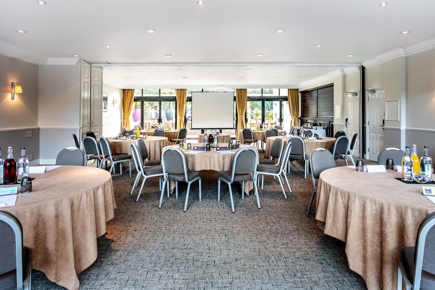 An event space with round tables covered in beige linens, grey chairs, and large windows at Pinewood on Wilmslow.