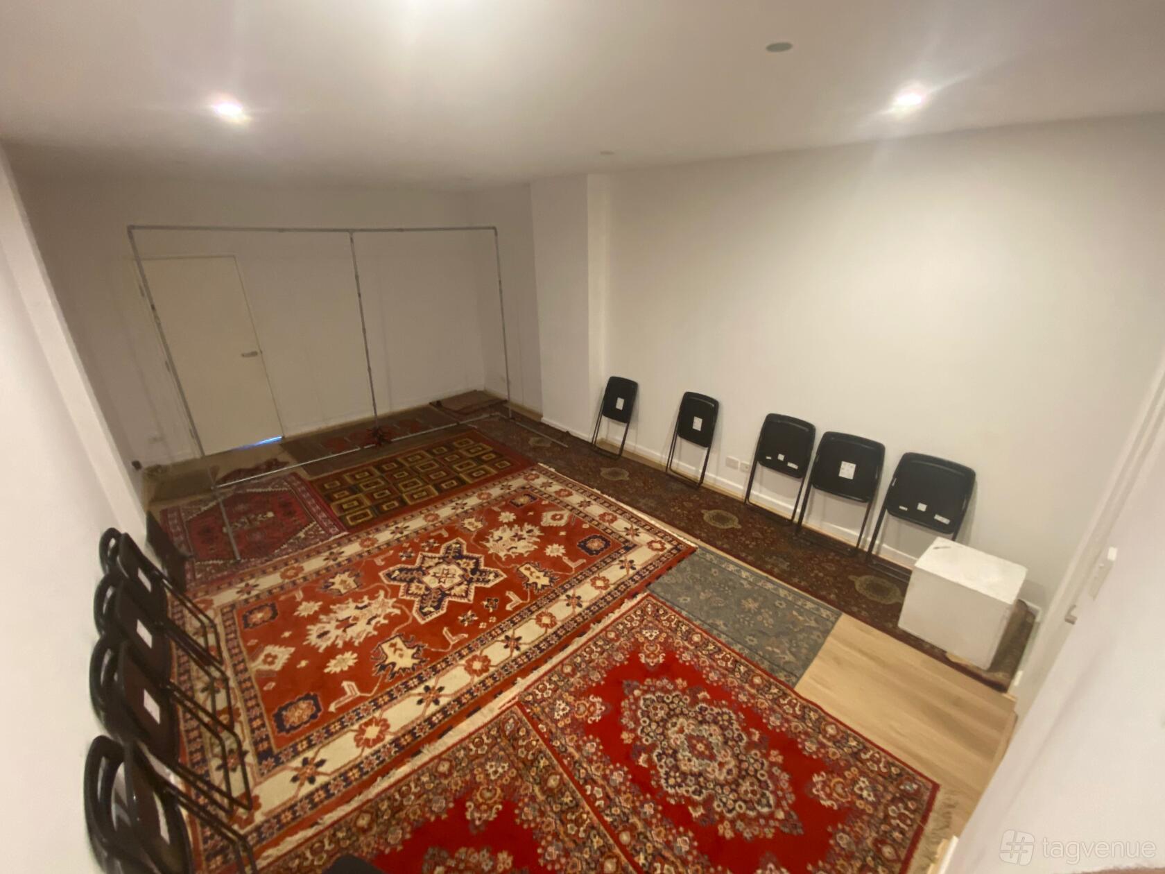 A photo studio with patterned rugs, white walls, and black folding chairs lined against the wall at Between Two Worlds Studio.