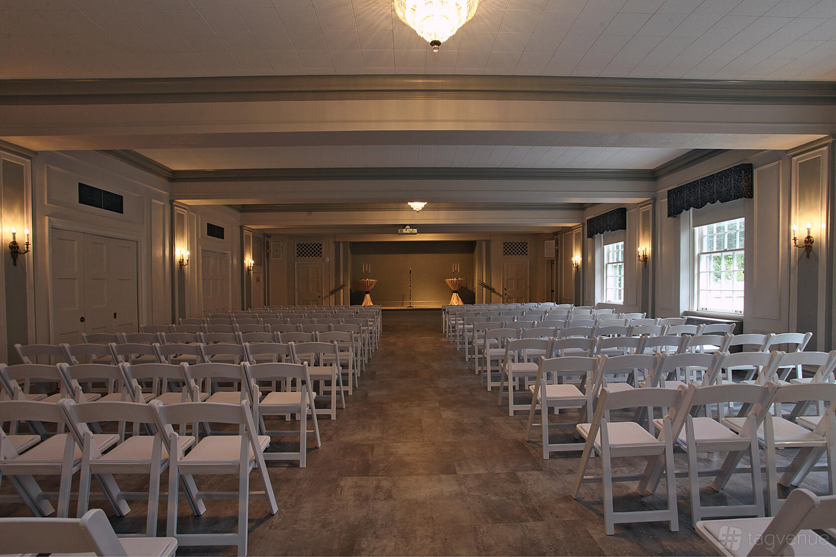 An event space with rows of white folding chairs facing a small stage and large windows at The Nineteenth Century Charitable Association.