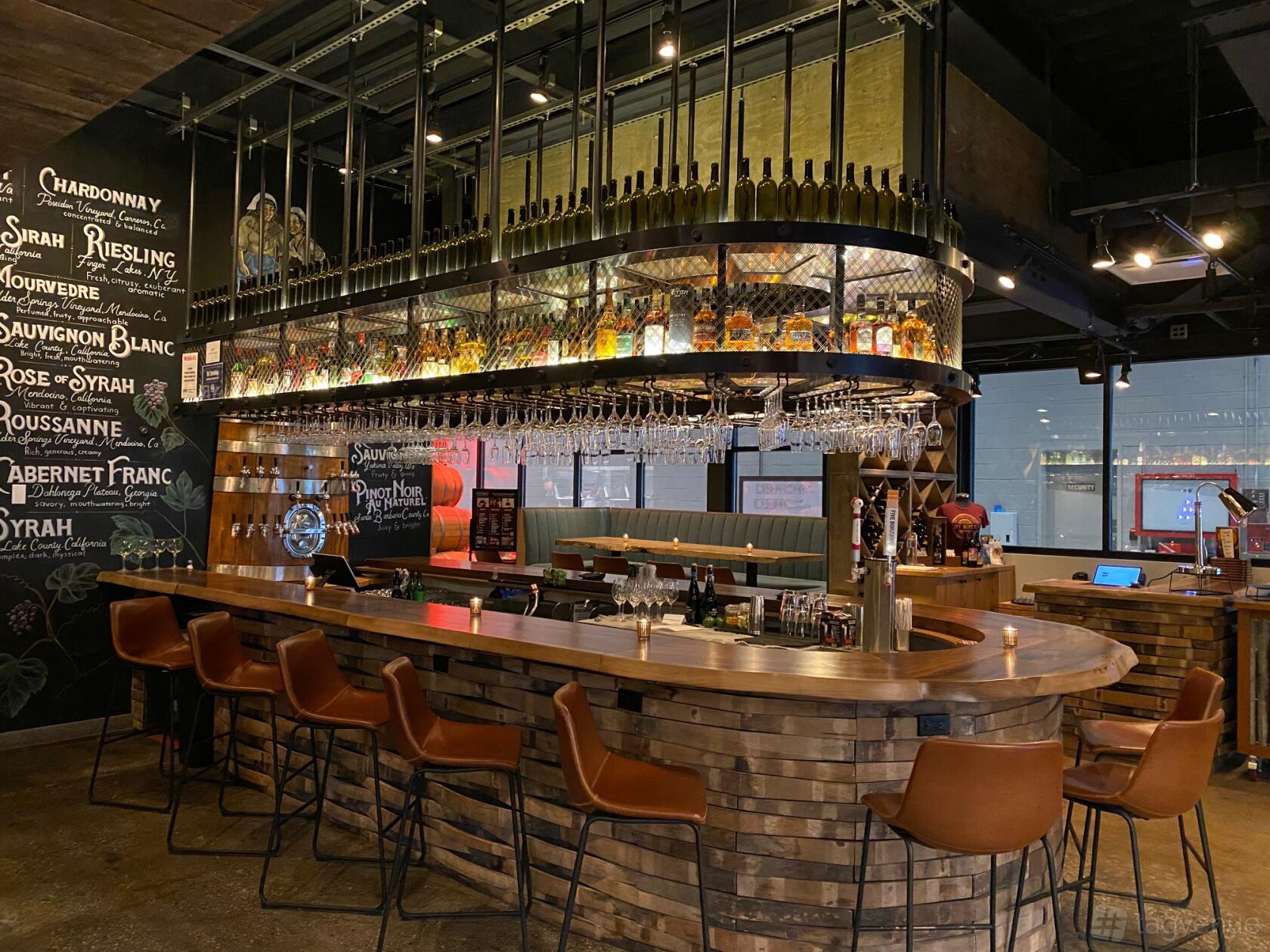A bar with a curved wooden counter, hanging glass racks, and backlit wine bottles at City Winery.