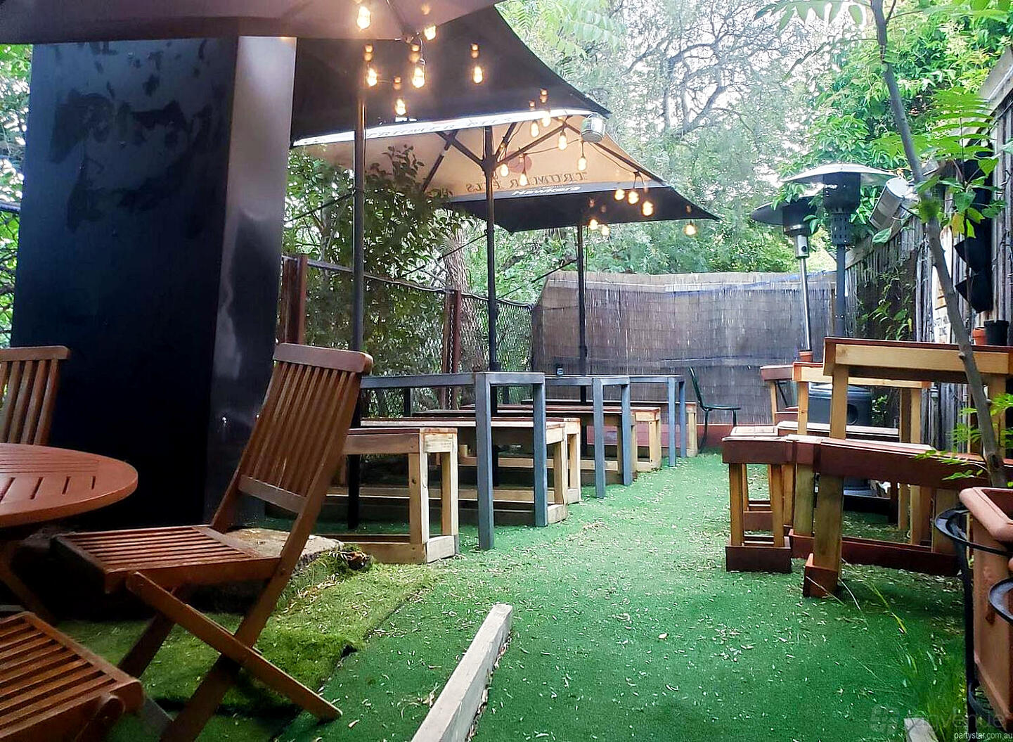 An outdoor space with wooden tables, string lights, umbrellas, and artificial grass at Radium Bar.