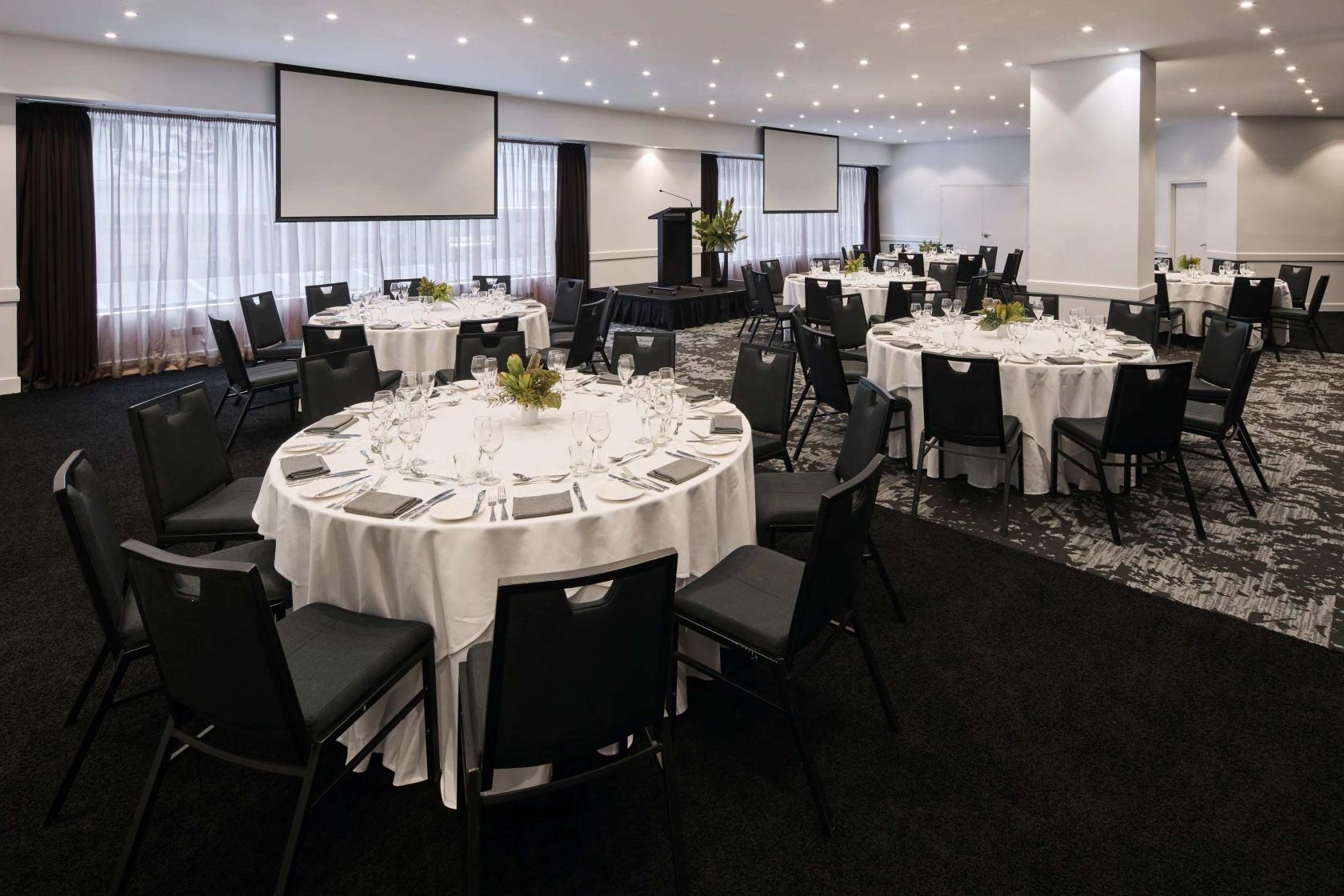 A hotel ballroom with round tables draped in white linens, black chairs, and two projection screens at Rydges World Square.