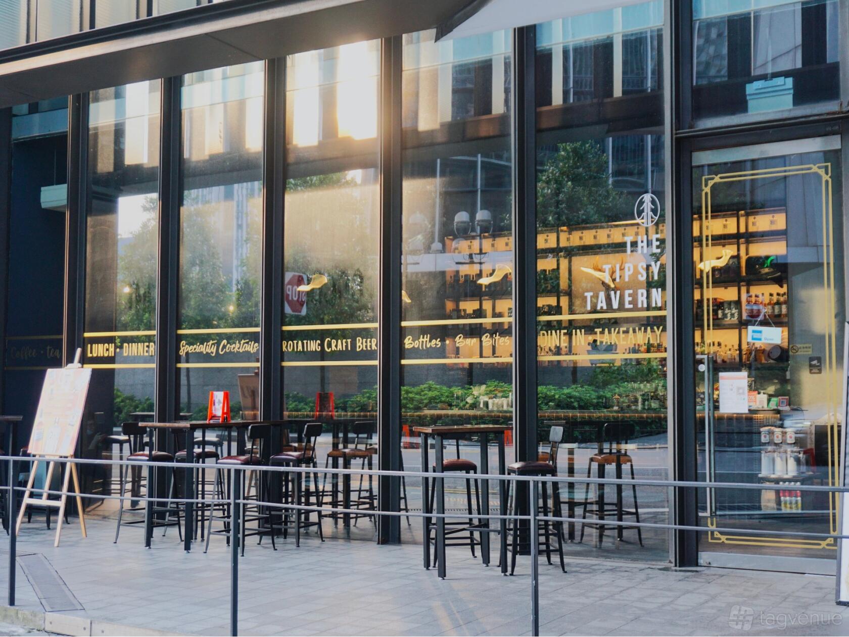 A pub with high tables and stools arranged on an outdoor patio in front of The Tipsy Tavern's glass facade.