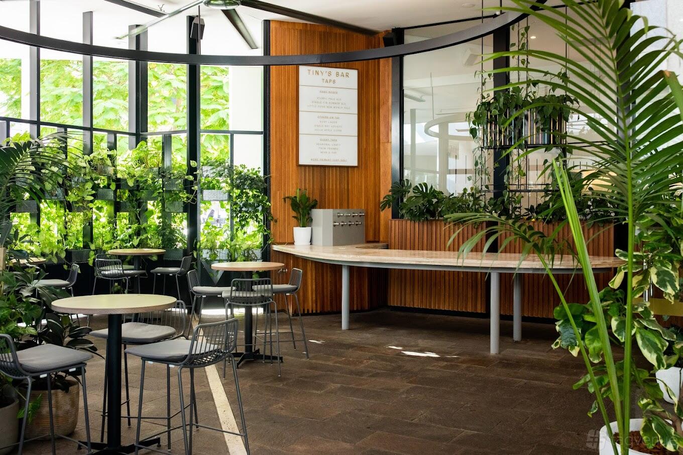An event space in a bar with potted plants, round tables, and floor-to-ceiling windows at Tiny's.