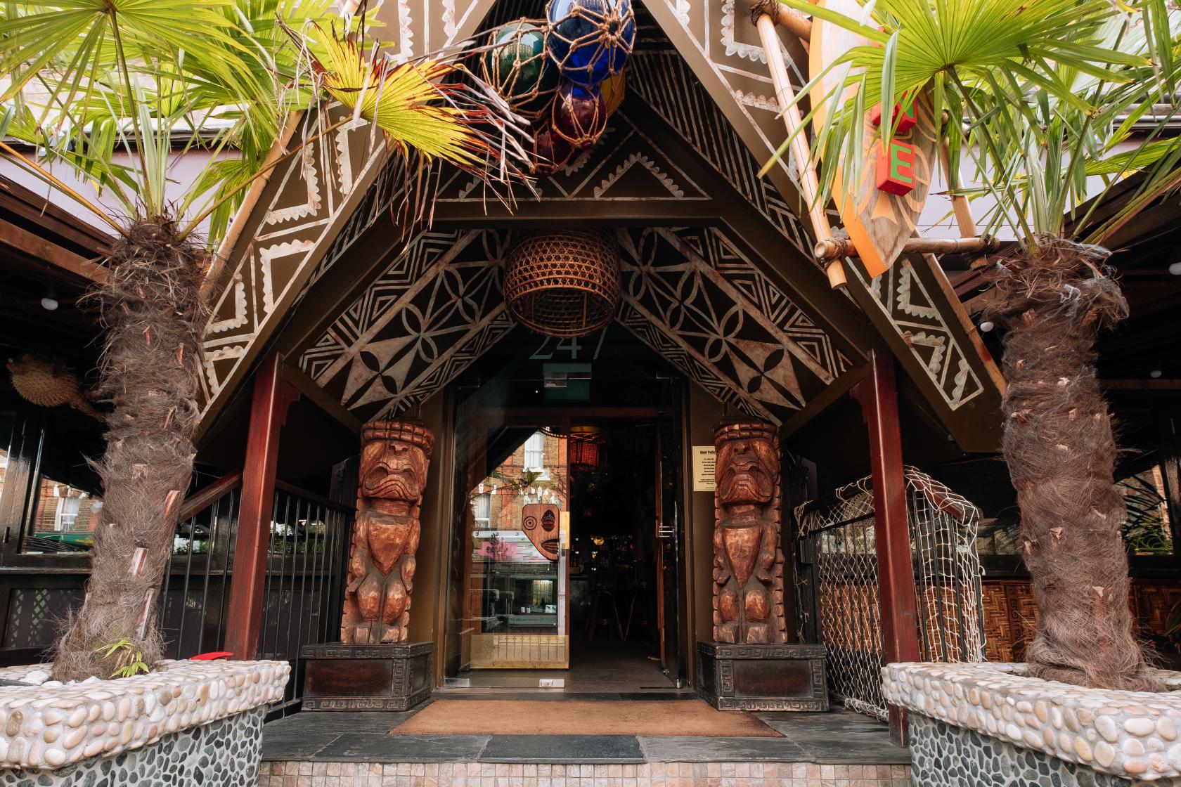 A tiki-inspired pub entrance with carved wooden statues, palm trees, and geometric patterned decor at Sugar Cane.