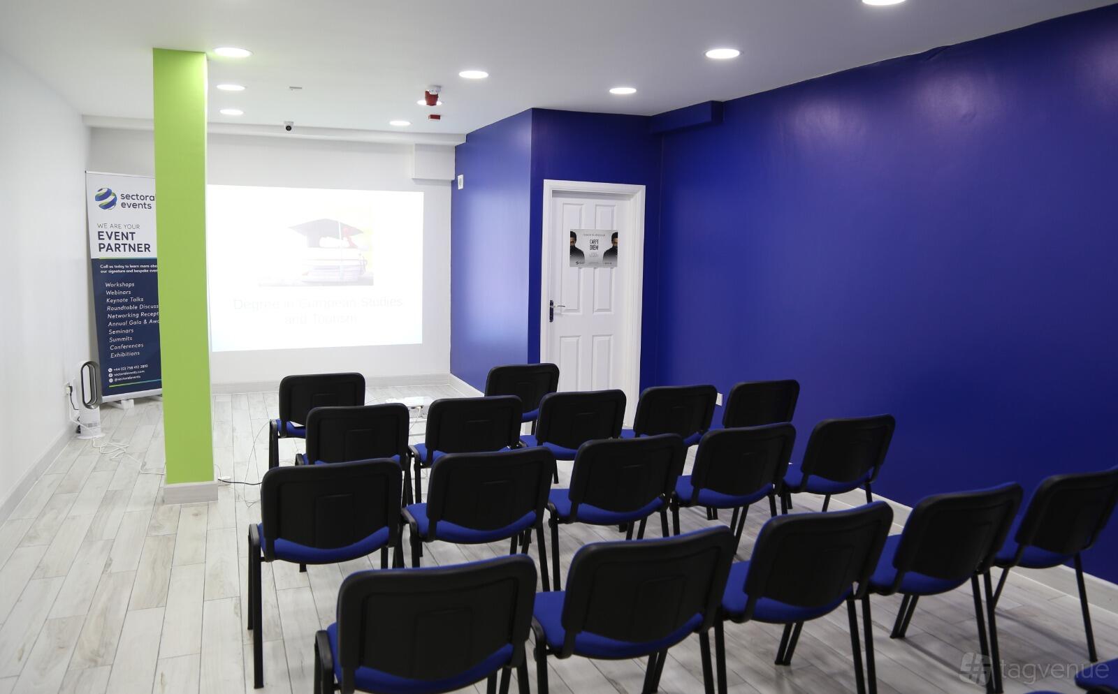 A meeting room with rows of black chairs facing a projector screen and walls painted blue and green at Edwin Holidays.
