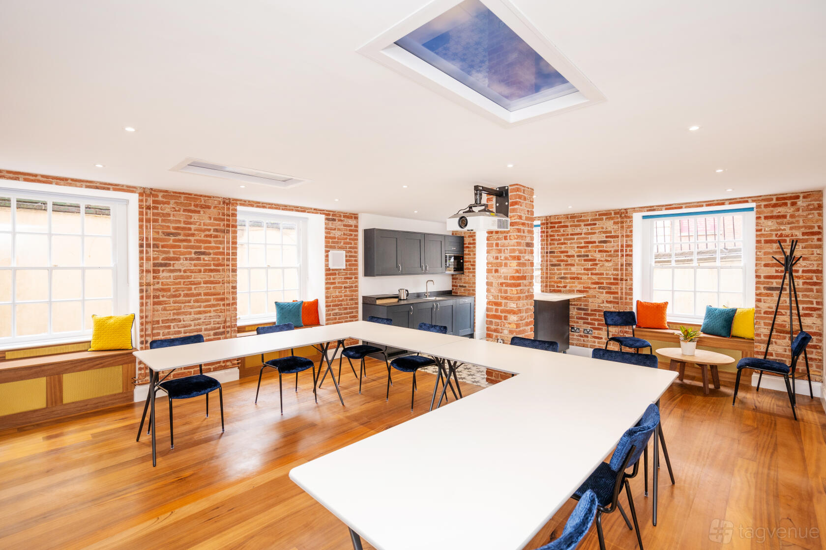 A meeting room with exposed brick walls, large windows, U-shaped tables, and a ceiling skylight at Square Works.