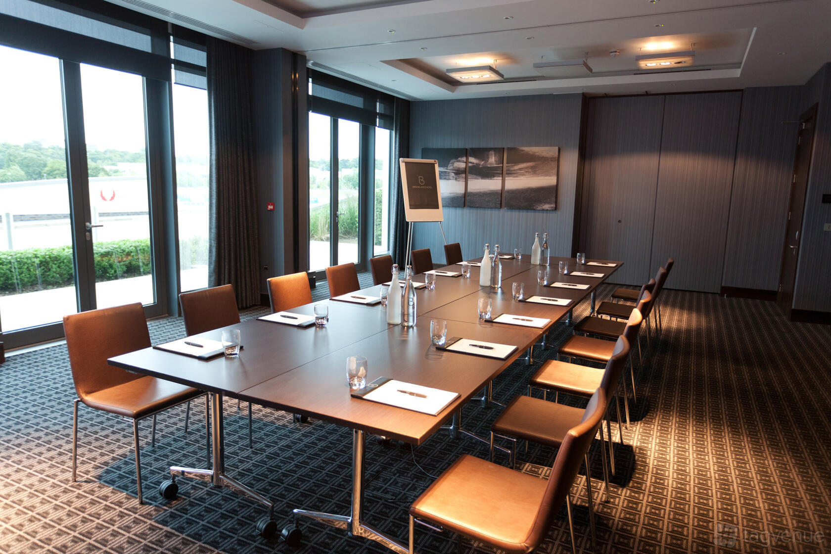 A meeting room with a large conference table, brown chairs, floor-to-ceiling windows, and notepads at Brooklands Hotel.