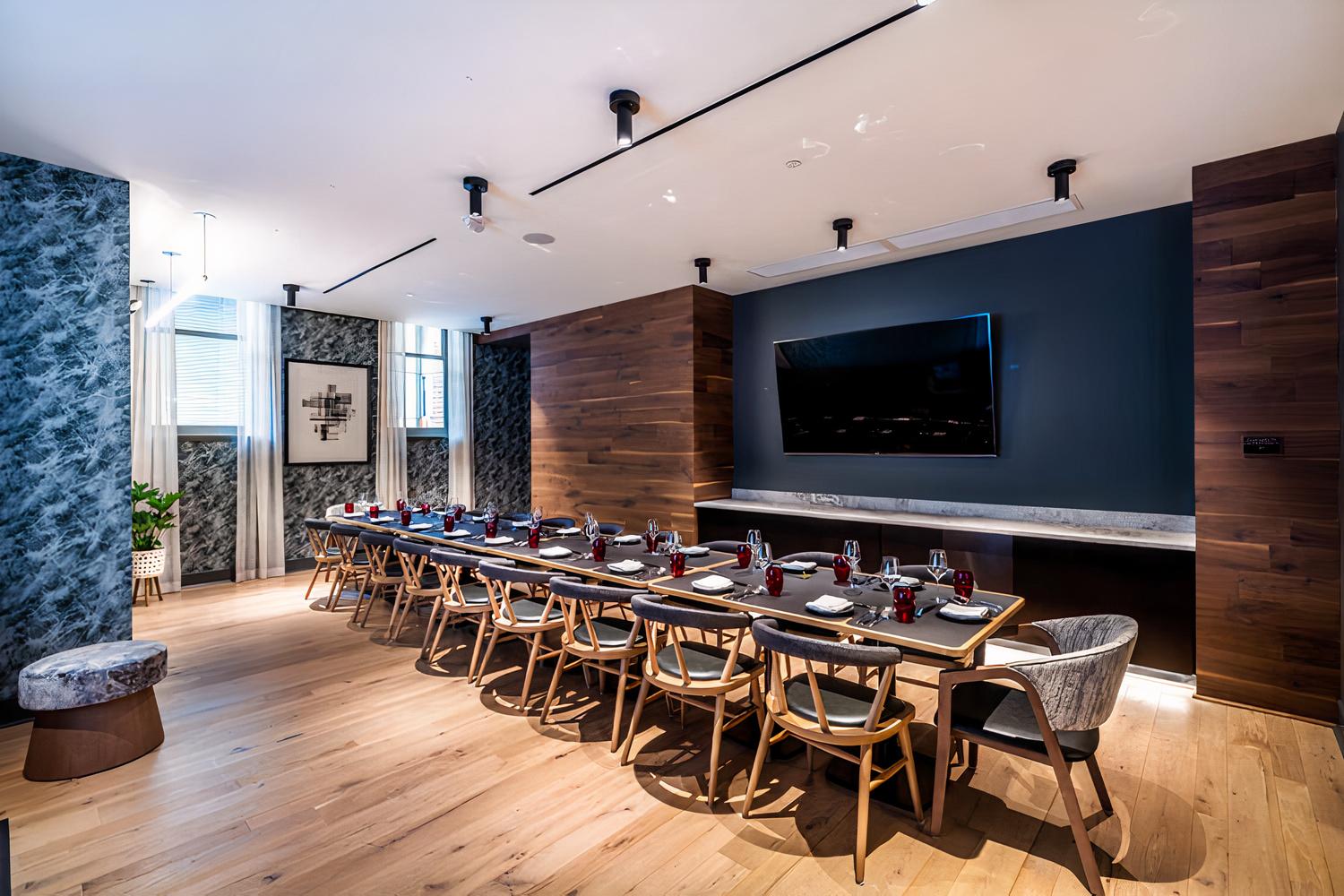A dining room with a long table set with black plates and red glasses, wood accents, and a large wall-mounted TV at Cranes Spanish Kaiseki.