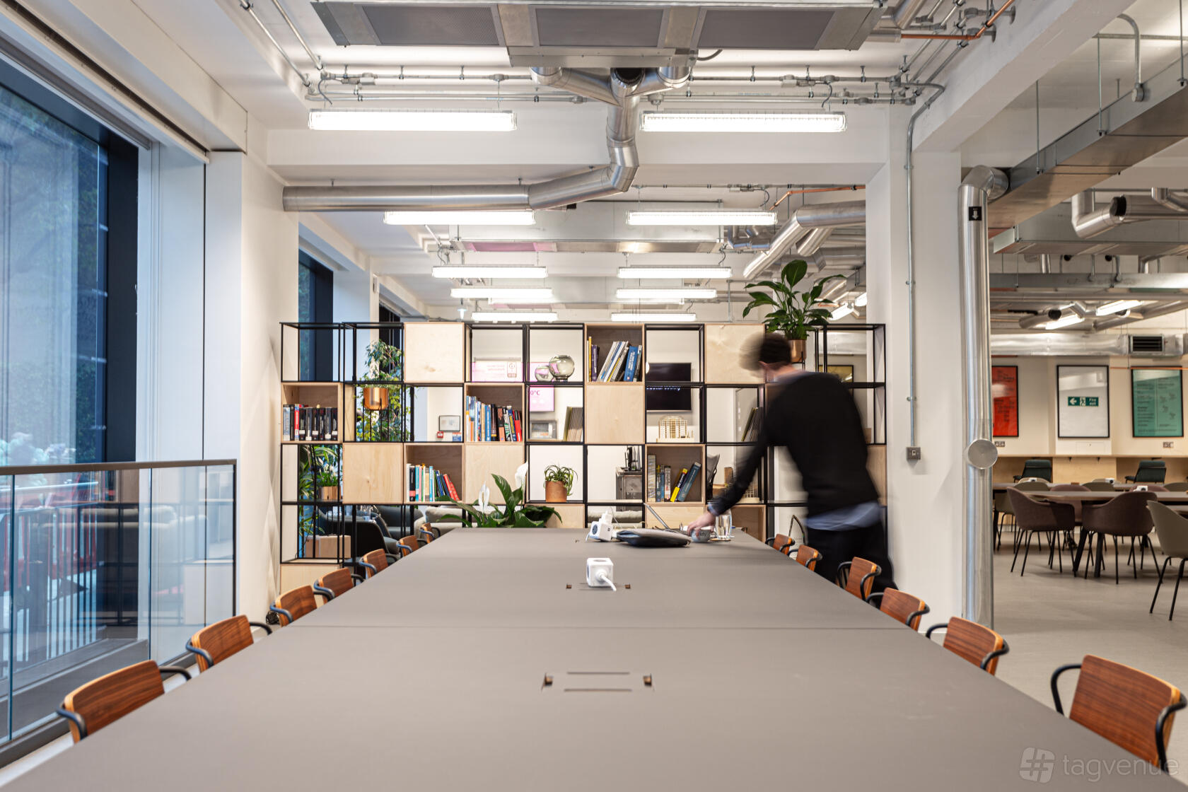 A meeting room with a long table, wooden chairs, open shelving with books, and large windows at The Society Building - Fitzrovia.
