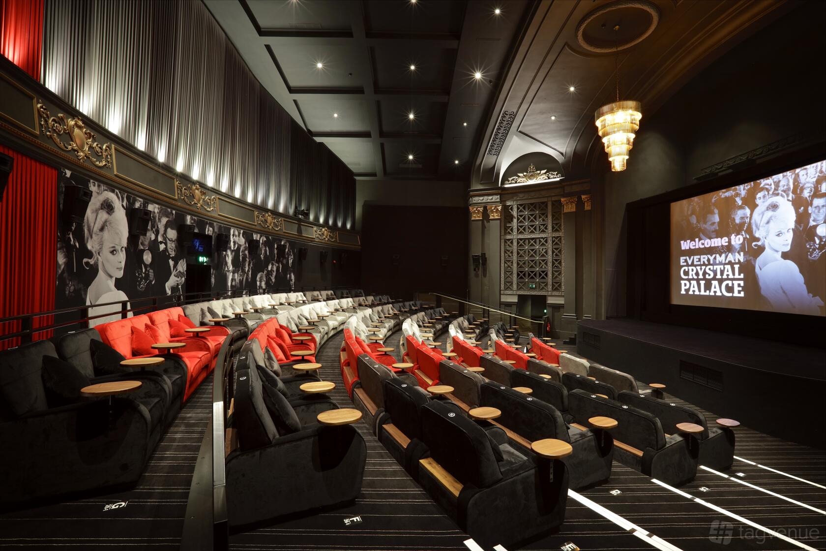An auditorium with tiered sofa seating, small tables, red accents, and chandeliers at Everyman Crystal Palace.