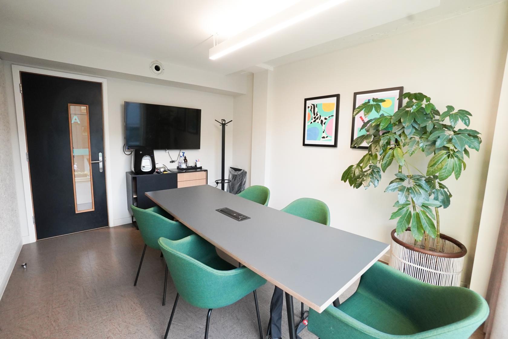 A meeting room with a rectangular table, green chairs, wall-mounted TV, and potted plant at The London Interdisciplinary School.