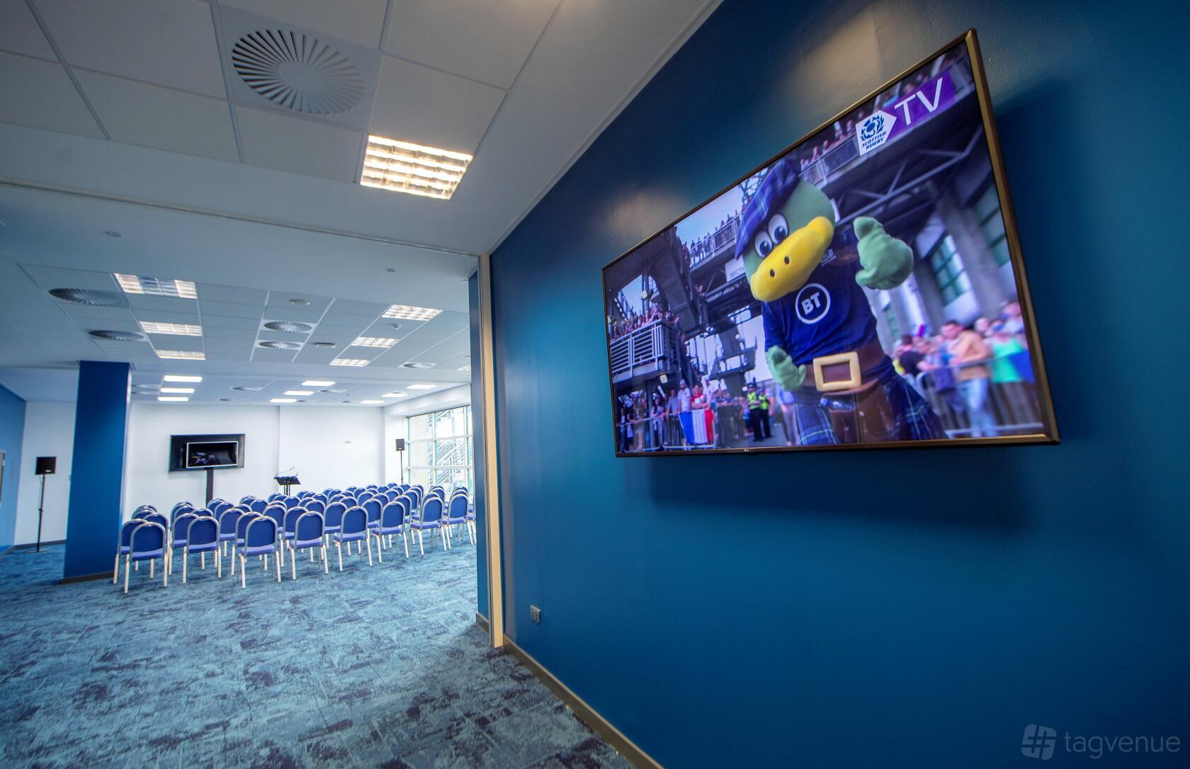 A stadium suite with rows of blue chairs, large windows, and a wall-mounted TV screen at BT Murrayfield Stadium.