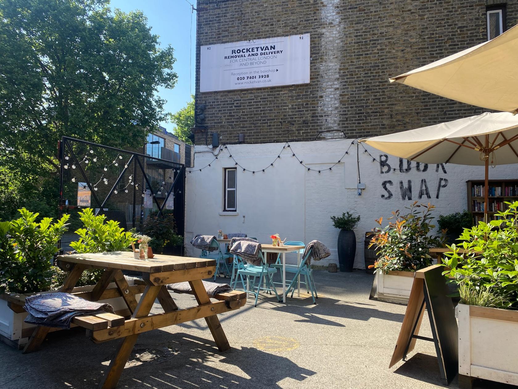 An outdoor cafe event space with picnic tables, potted plants, and bookshelves at Rocketvan.