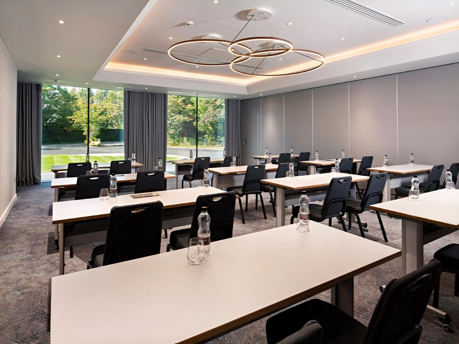 A meeting room with large windows, modern light fixtures, and rows of tables with bottled water at Holiday Inn Shepperton.