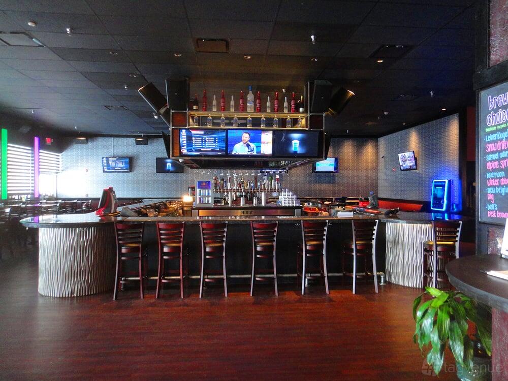 A bar with a curved counter, high chairs, wall-mounted TVs, and decorative bottle display at House of Music & Entertainment.