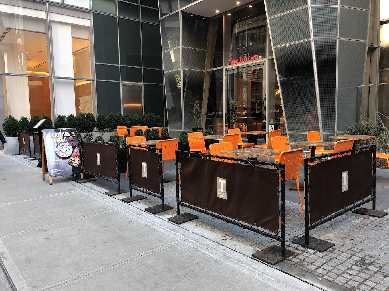 An outdoor dining area with orange chairs, black barricades, and potted plants at Tarallucci e Vino NoMad.
