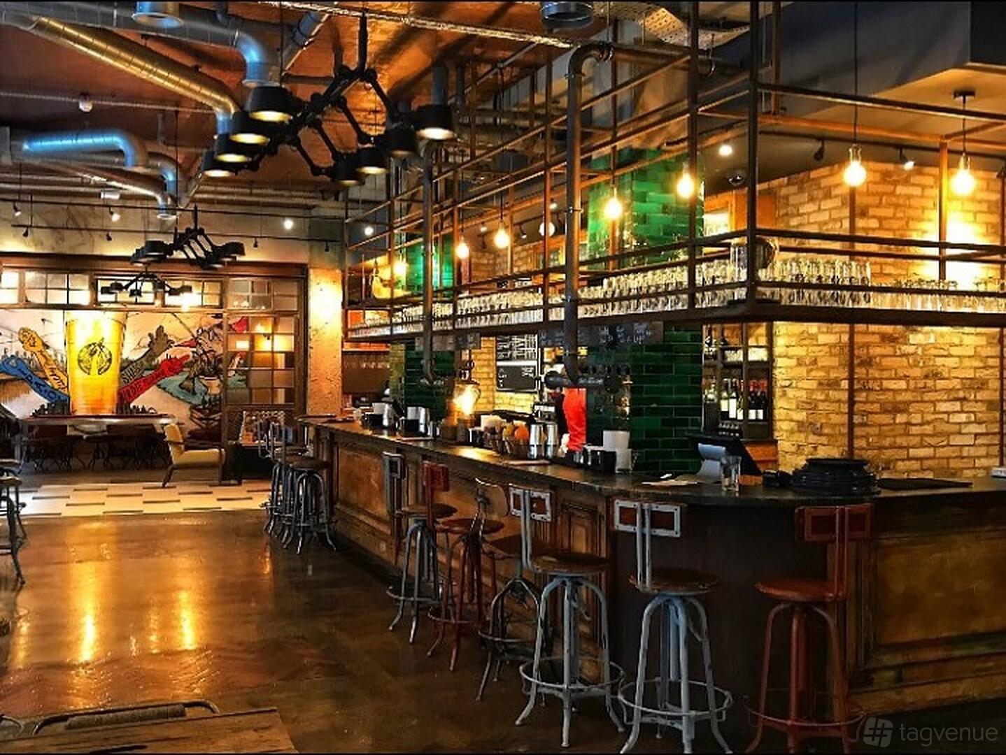 A pub with exposed brick walls, industrial lighting, and a long bar lined with metal stools at Long Arm Brewery & Tap Room.