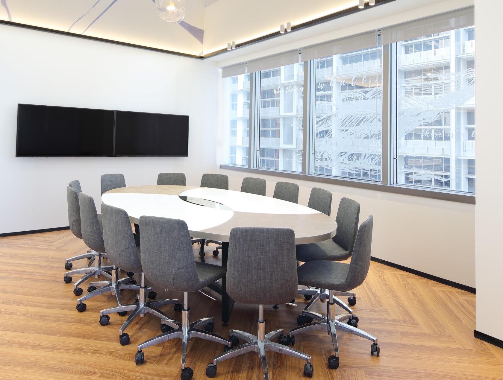 A meeting room with a large oval table, twelve gray chairs, two wall-mounted screens, and floor-to-ceiling windows at The Hive Tanjong Pagar.