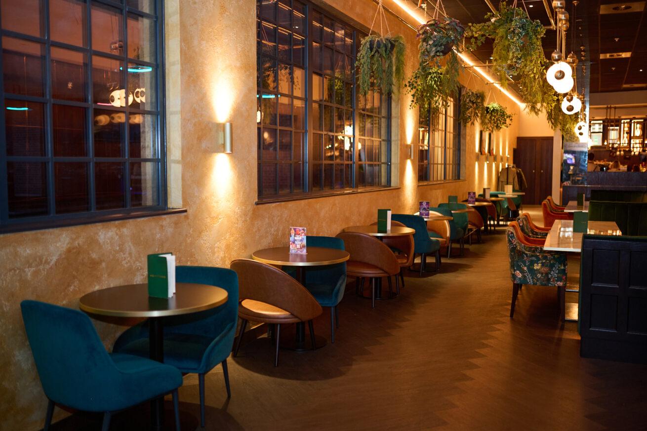 A bar with velvet chairs, round tables, large industrial windows, and hanging greenery at Gravity - Wandsworth.
