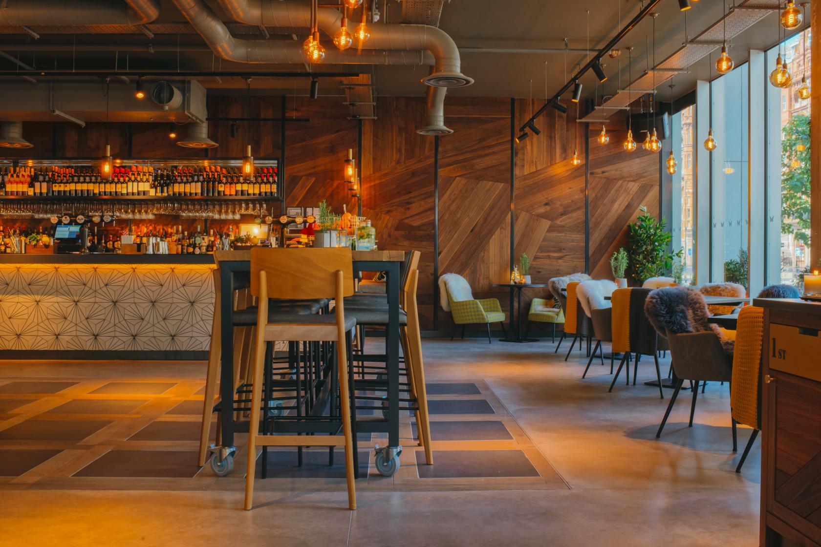A cocktail bar with geometric wood paneling, hanging Edison bulbs, and floor-to-ceiling windows at The Anthologist Manchester.