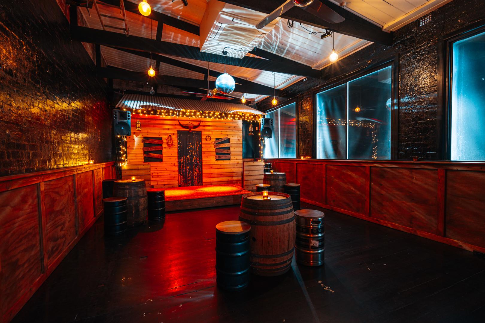 A function room with a small wooden stage, string lights, and barrel tables at Death or Glory.