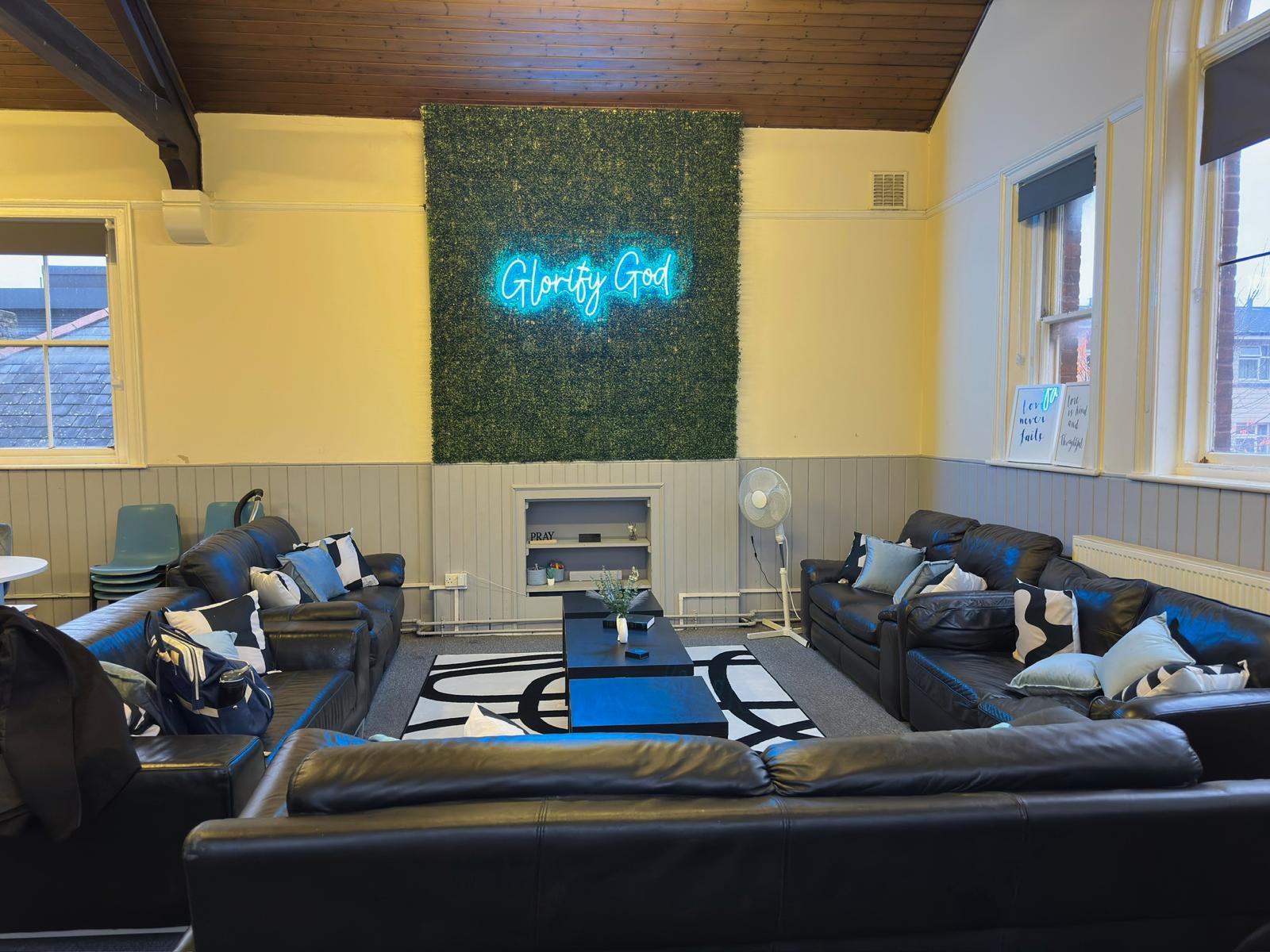 A function room with black leather sofas, a neon 'Glorify God' sign, and a patterned rug at West Croydon Baptist Church.