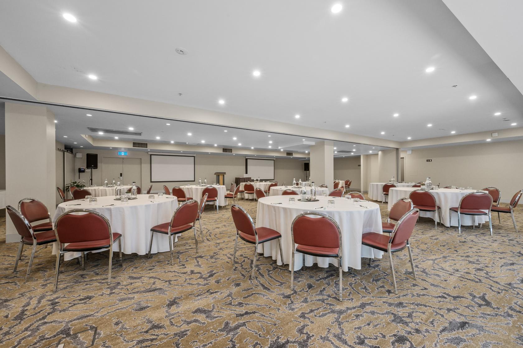 An event space with round tables covered in white linens, red chairs, and ceiling spotlights at Batman's Hill on Collins.