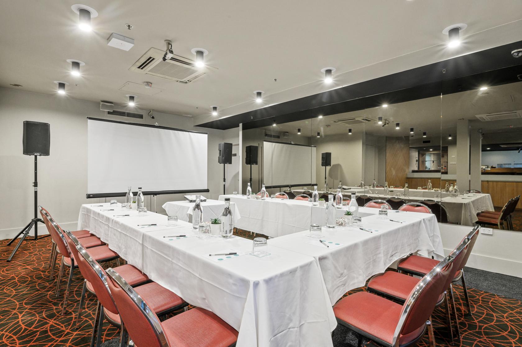 A hotel meeting room with U-shaped tables covered in white linens, red chairs, and a large projection screen at Batman's Hill on Collins.