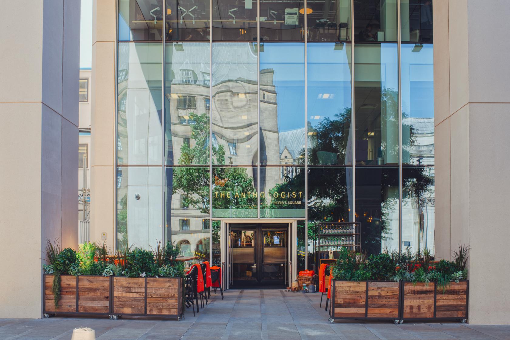 A bar with tall glass windows, planters with greenery, and outdoor seating at The Anthologist Manchester.