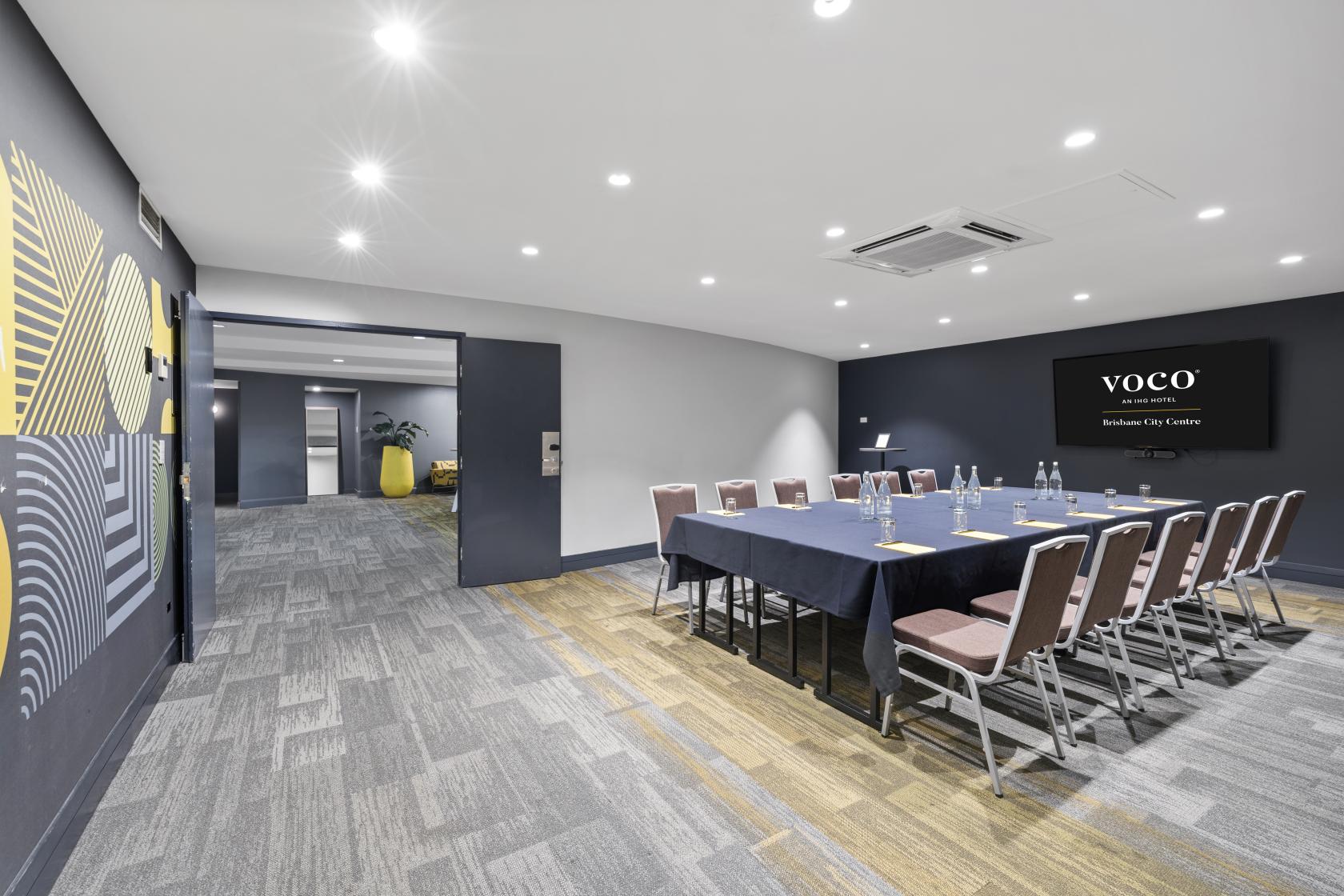 A hotel meeting room with a U-shaped table arrangement, wall-mounted screen, and patterned carpet at Voco Hotel Brisbane City Centre.