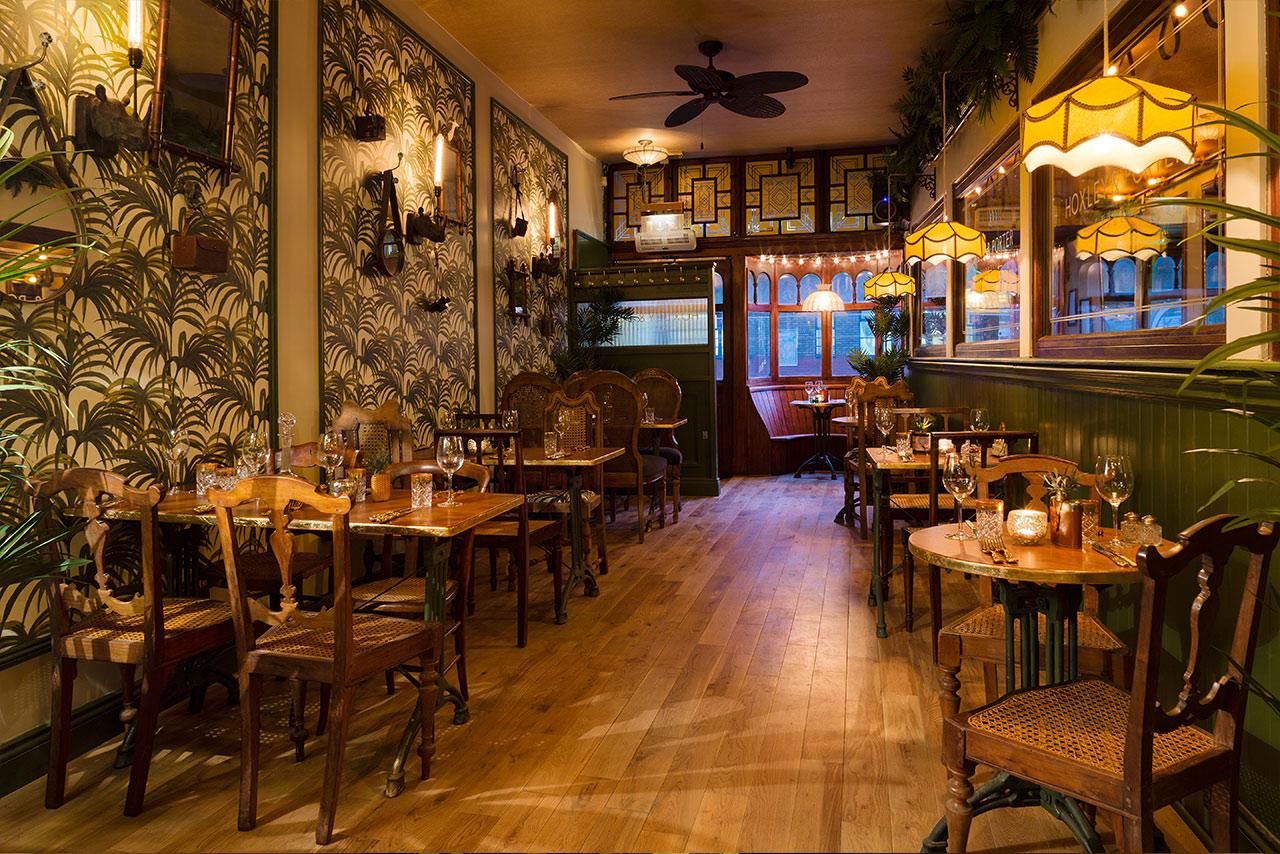 A dining room with patterned wallpaper, wooden tables, vintage chairs, and warm pendant lighting at Hoxley and Porter.