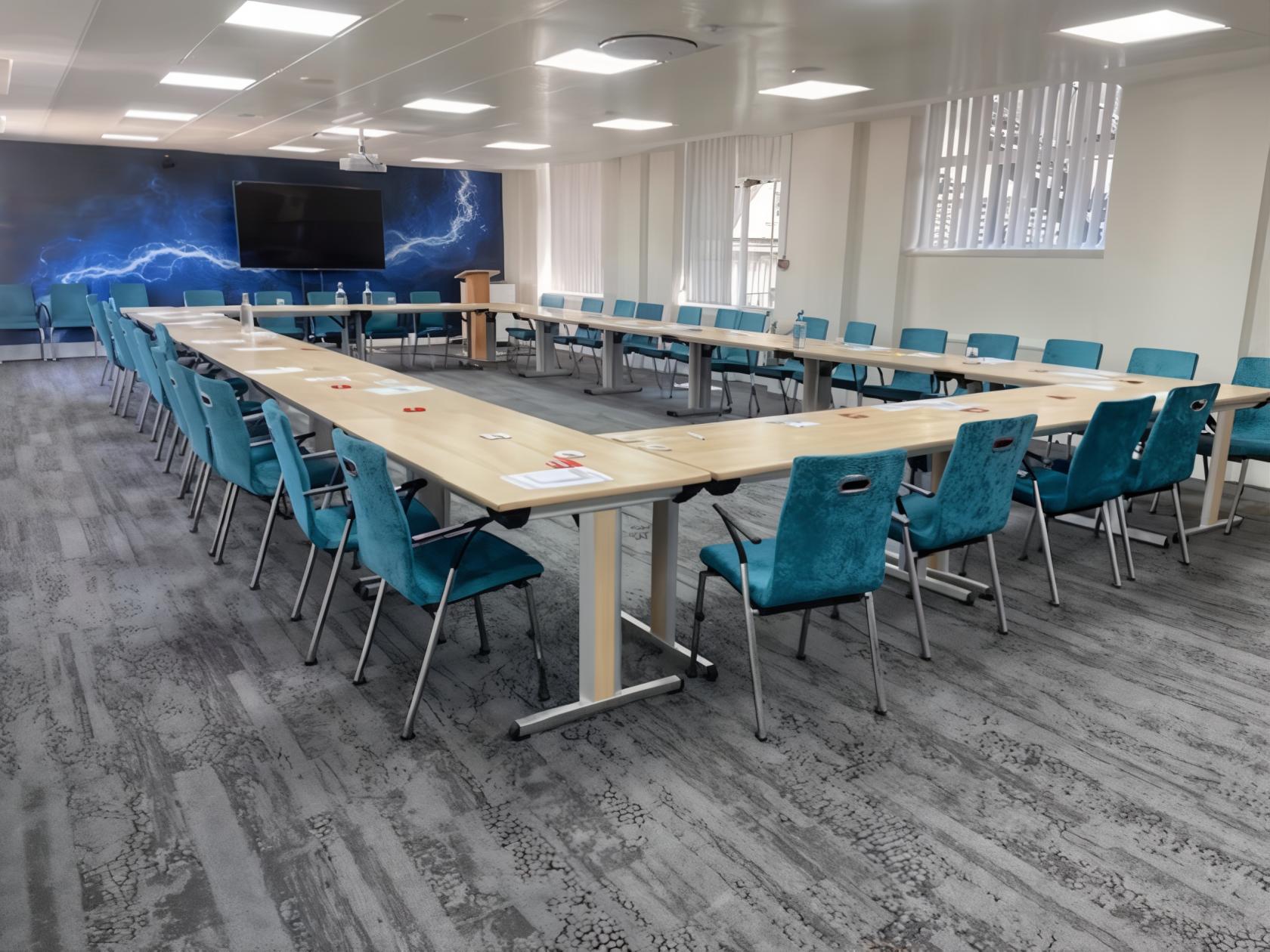 A meeting room with U-shaped tables, teal chairs, wall art with lightning, and a large screen at Broadway House.