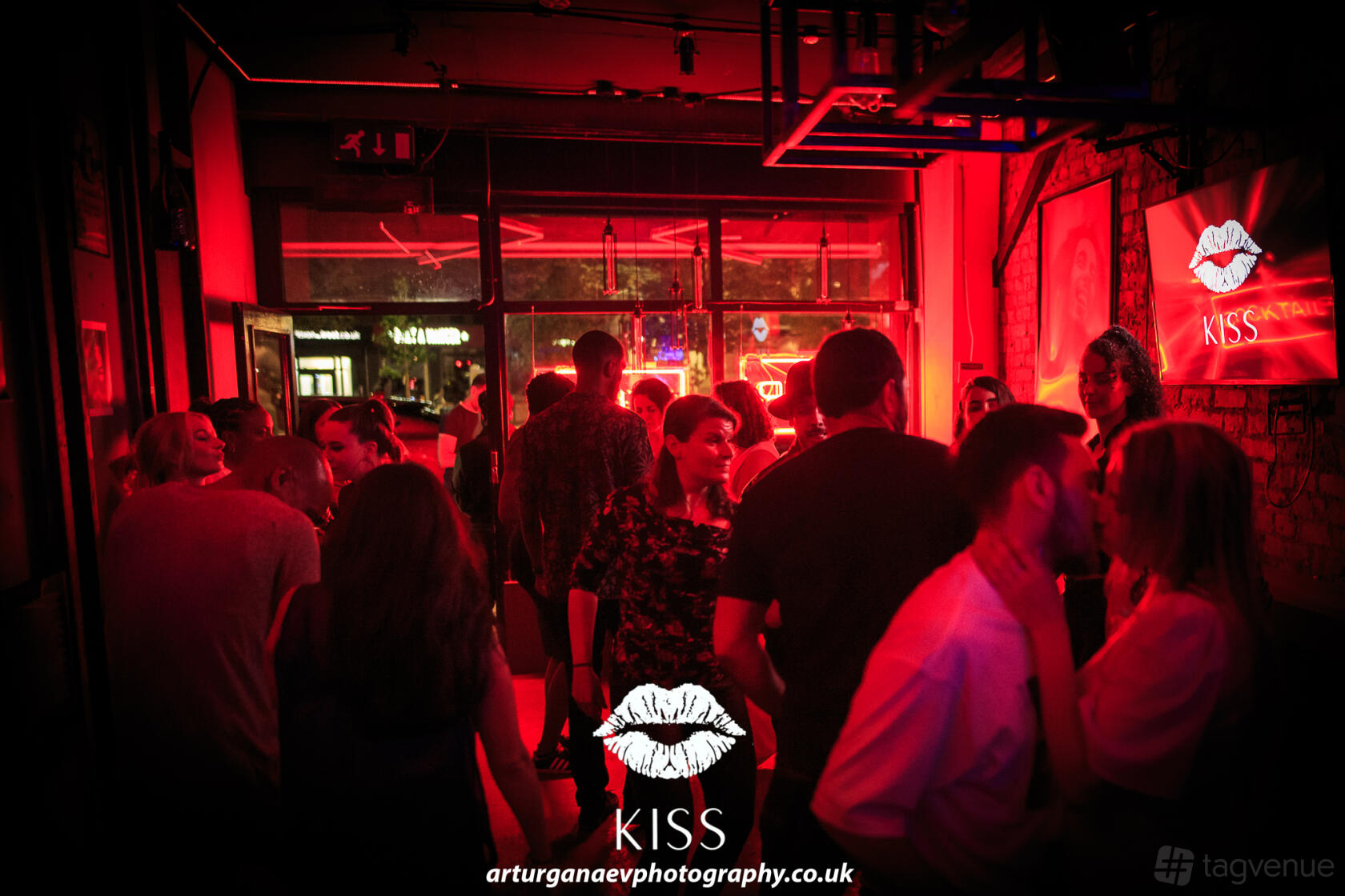 A lively bar with red neon lighting, exposed brick walls, and groups of people socializing at Kiss.