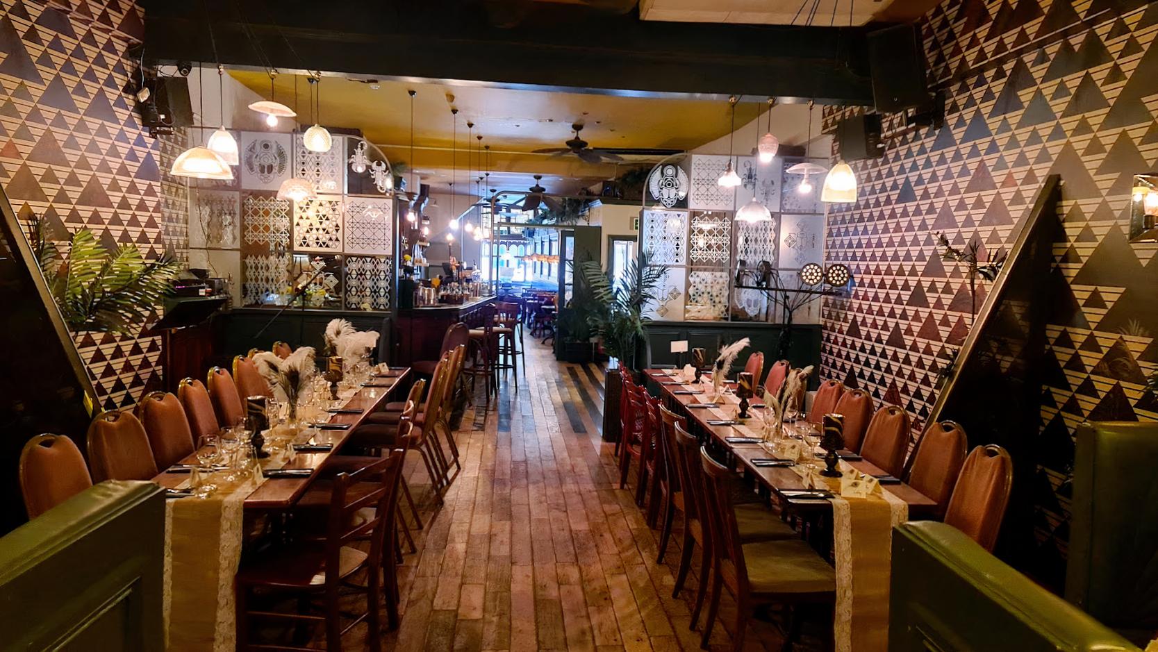 A dining room with patterned walls, pendant lighting, and long banquet tables set with glassware at Hoxley and Porter.