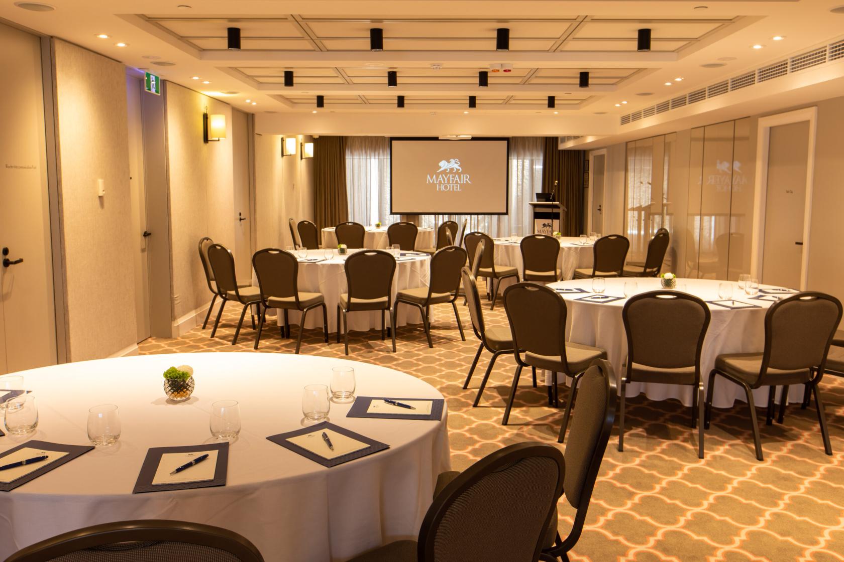 A function room with round tables set with glasses and notepads, patterned carpet, and a projector screen at Mayfair Hotel Adelaide.