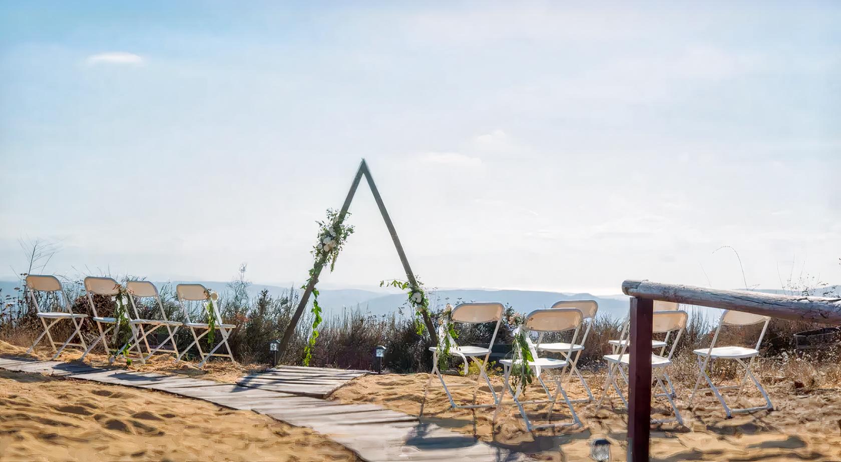 An outdoor ceremony area with folding chairs arranged on sand and a wooden triangular arch at Poway Equestrian Center.