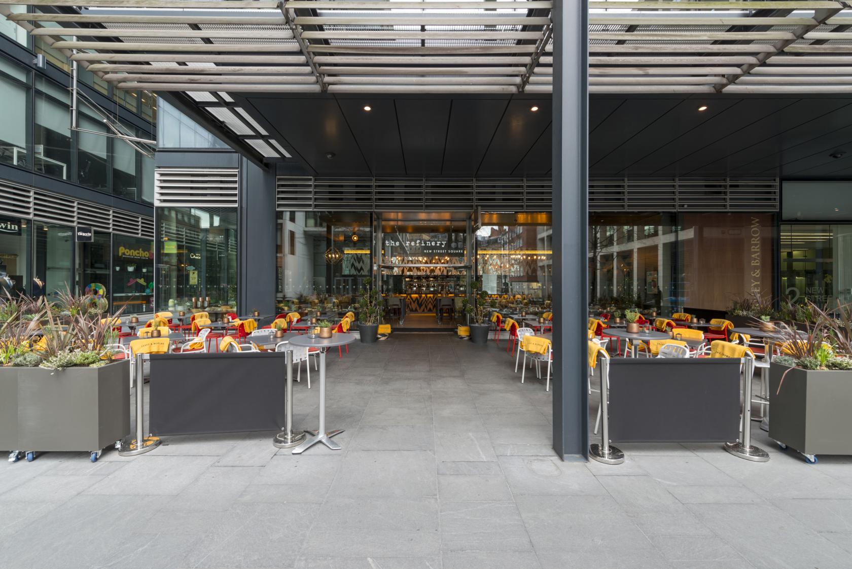 An outdoor cocktail bar in a restaurant with colorful chairs, large planters, and a covered patio at The Refinery New Street Square.