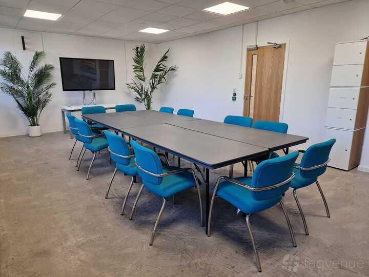 A boardroom in a coworking space with blue chairs, large tables, indoor plants, and a wall-mounted screen at MAKE IT Barking.