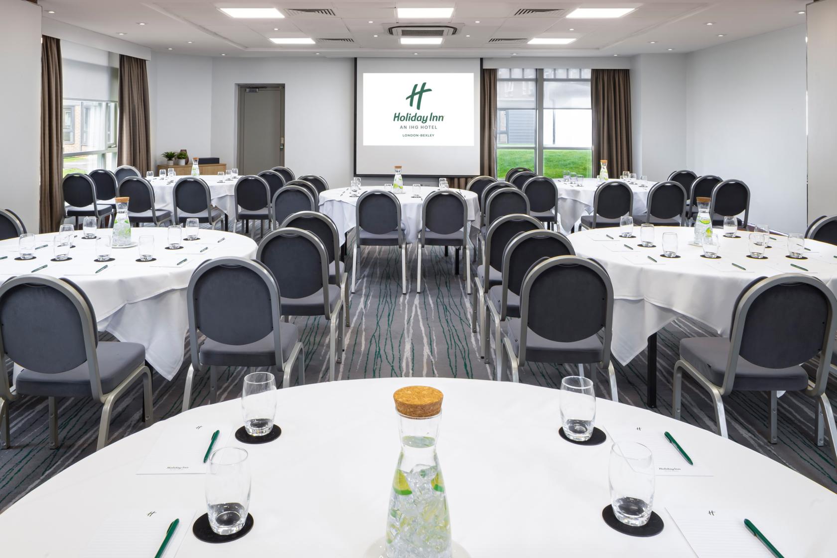 An event space with round tables, grey chairs, water pitchers, and a projector screen at Holiday Inn London Bexley.